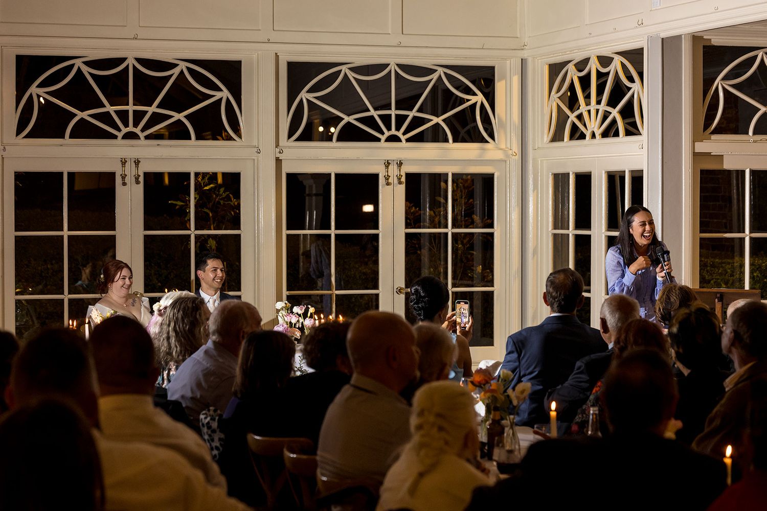 The Grooms Sister during speeches in an elegant room with decorative arched windows at night at Gabbinbar during a wedding reception.
