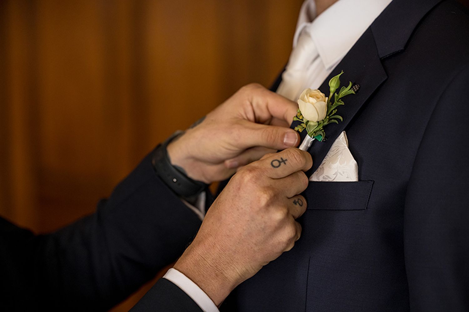 Hands pinning a cream rose and greenery boutonniere onto a navy suit lapel during wedding preparations at Gabbinbar Homestead in Toowoomba.