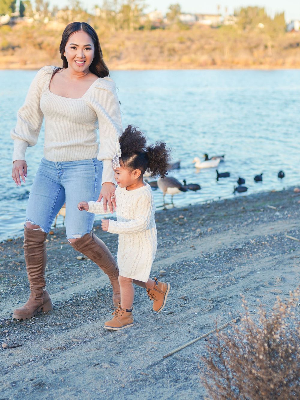 Parent and child in matching cream sweaters running together along a lakeshore with ducks in background.