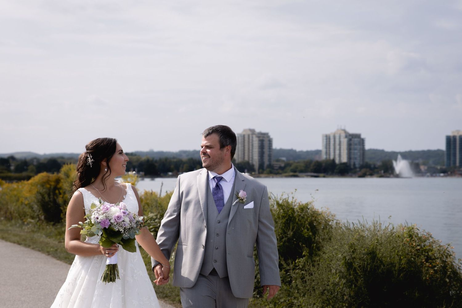 A newlywed couple walks along a waterfront path with city buildings and trees visible in the background on a cloudy day.