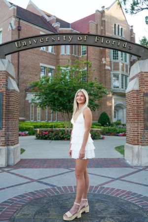 Graduate poses under the University of Florida entrance arch in a white dress and heeled sandals.