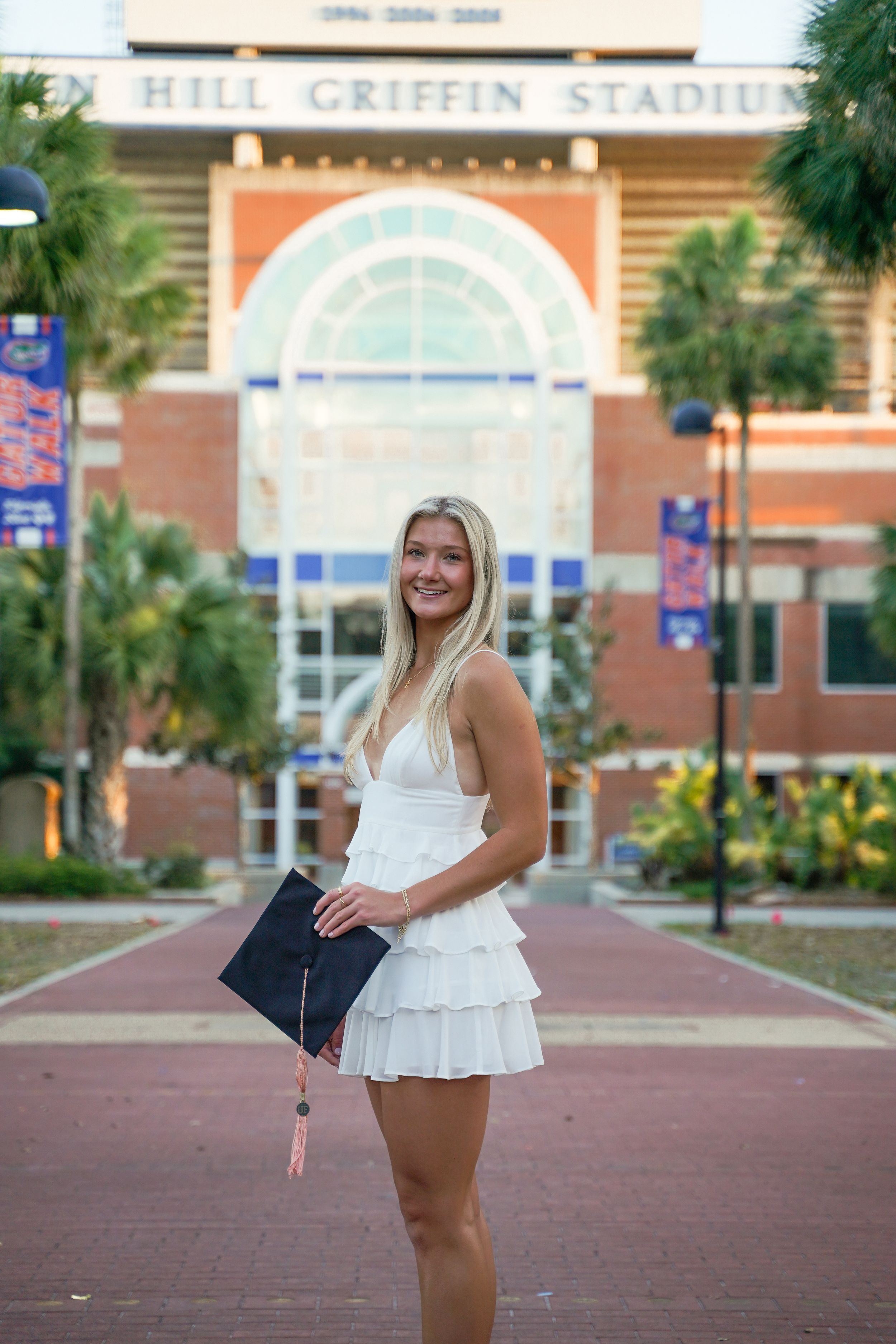 Graduate stands in front of Ben Hill Griffin Stadium wearing a white dress and holding a diploma.