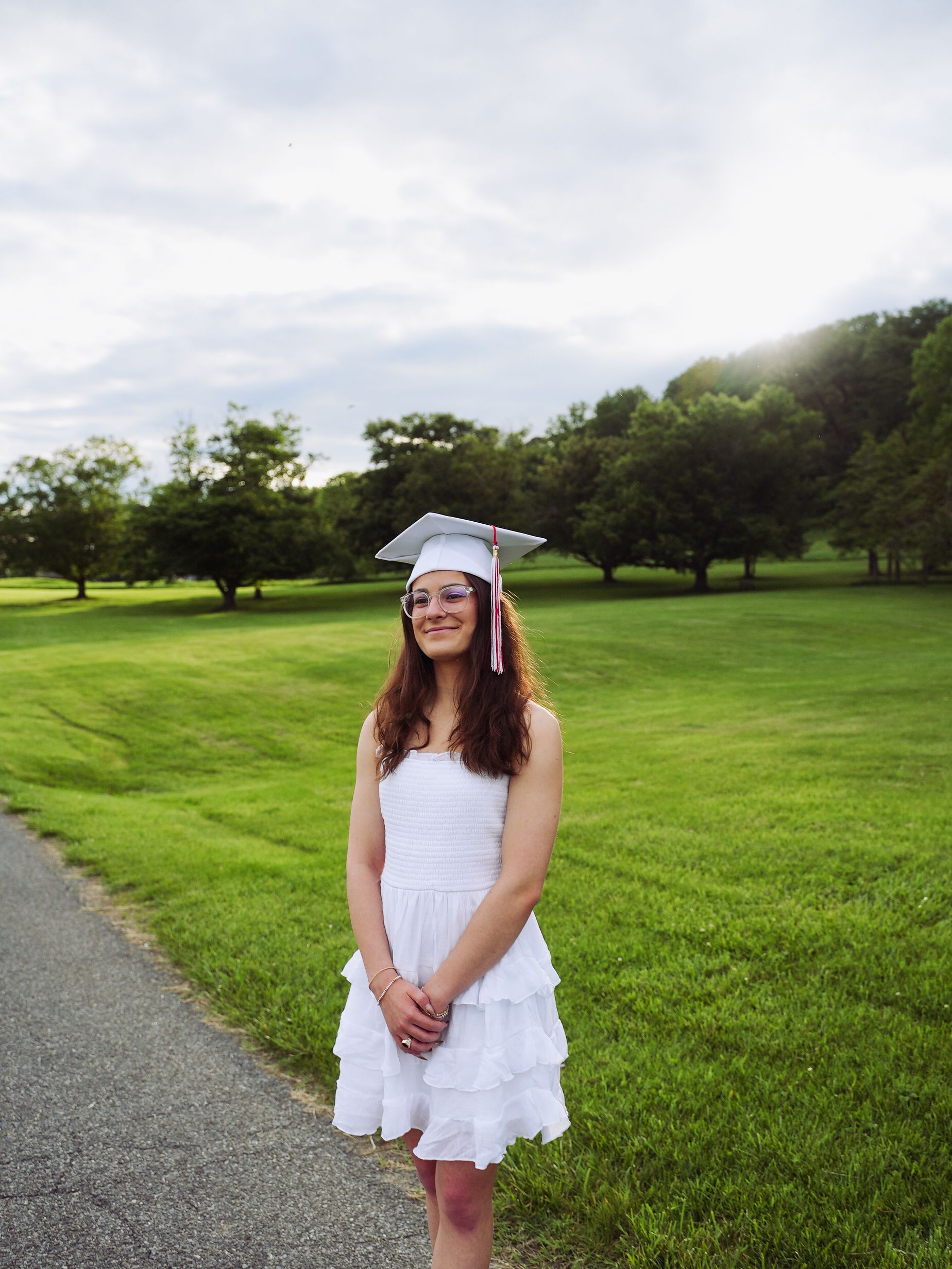 Graduate in white dress and cap poses on path with lush green lawn and trees in background on sunny day.