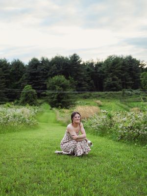 A person in a floral dress sits peacefully on a grassy lawn surrounded by evergreen trees and wildflowers.