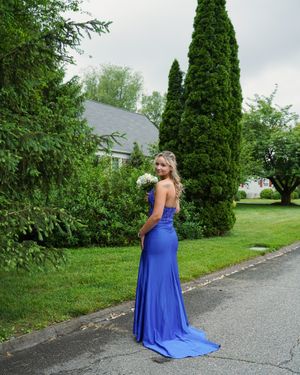 A person in a royal blue formal gown holds flowers in a garden setting.