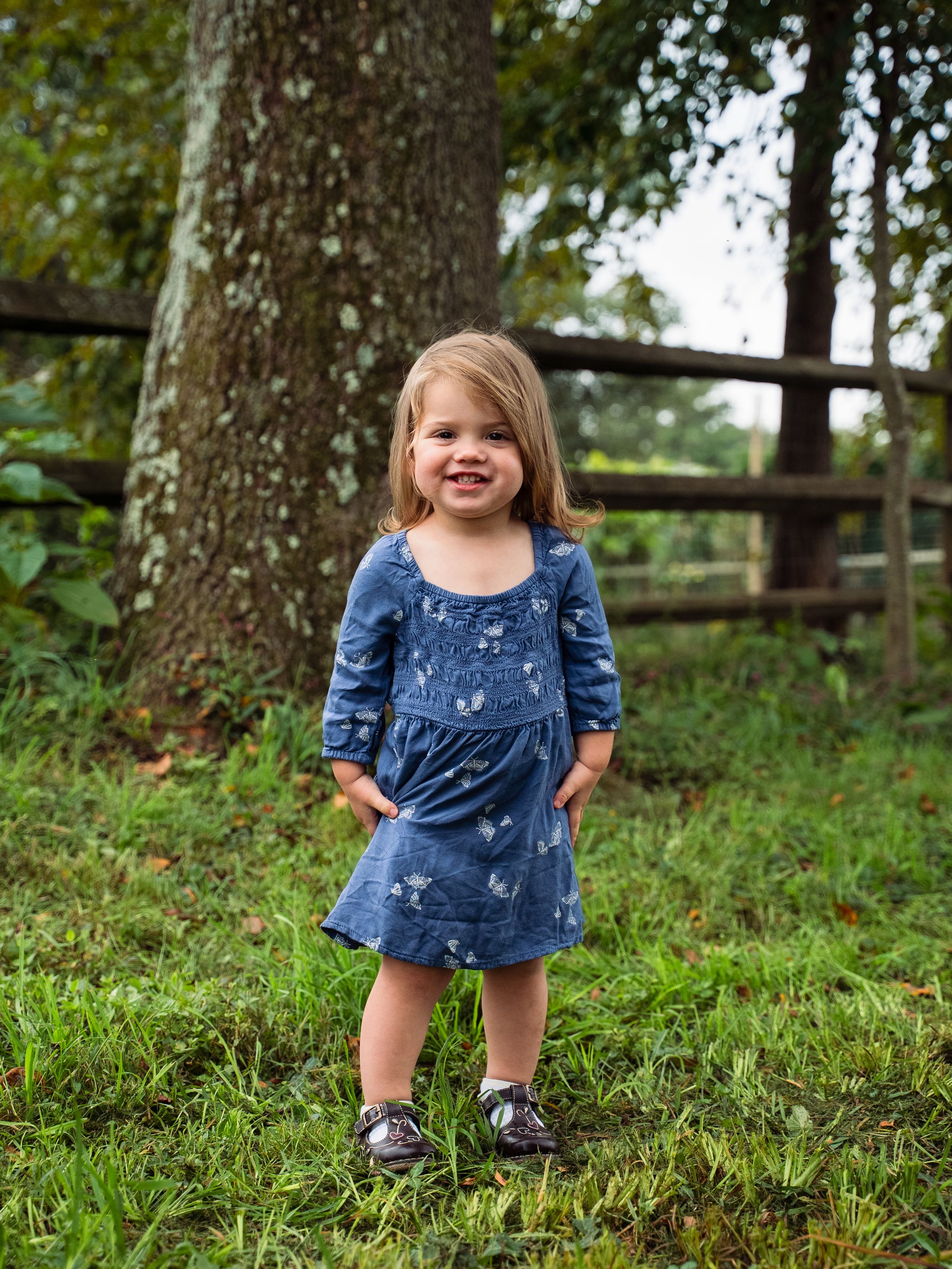 A series of candid photos showing a toddler in a blue dress playing and laughing on grassy ground near trees.