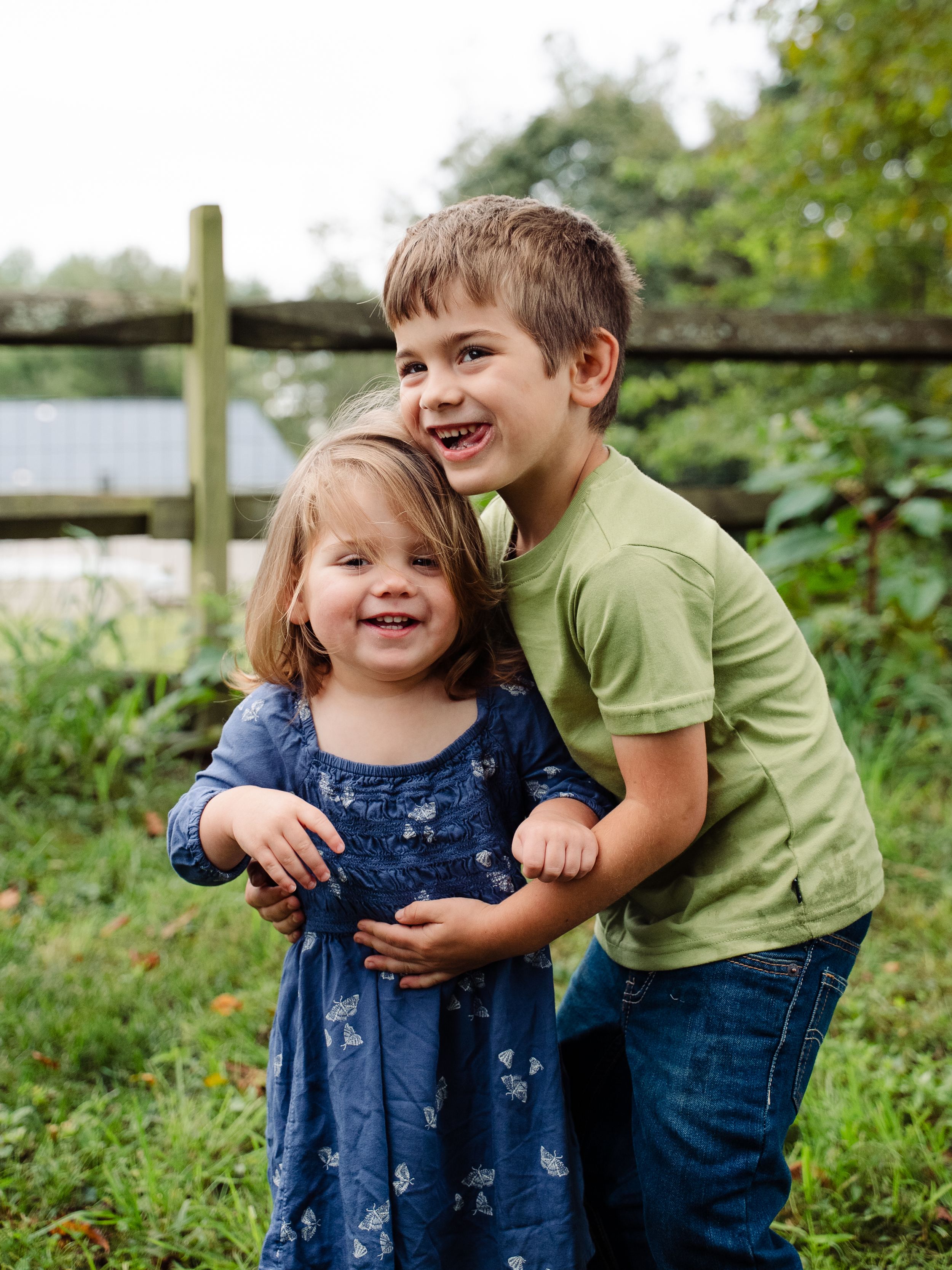 Two children share a sweet moment outdoors in a garden setting, one wearing a blue dress and one in green.