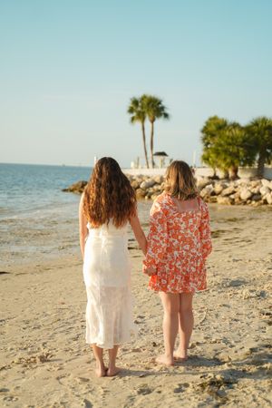Two people in dresses walking together on a beach with palm trees and rocks in the background.