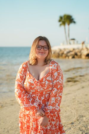 A person wearing a vibrant orange floral dress poses on a beach with palm trees in the background.