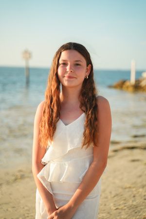 A person in a white dress poses on a sunny beach with palm trees and ocean in the background.