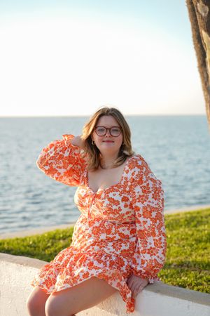 A person in an orange floral dress sits on a white wall by the ocean during golden hour.
