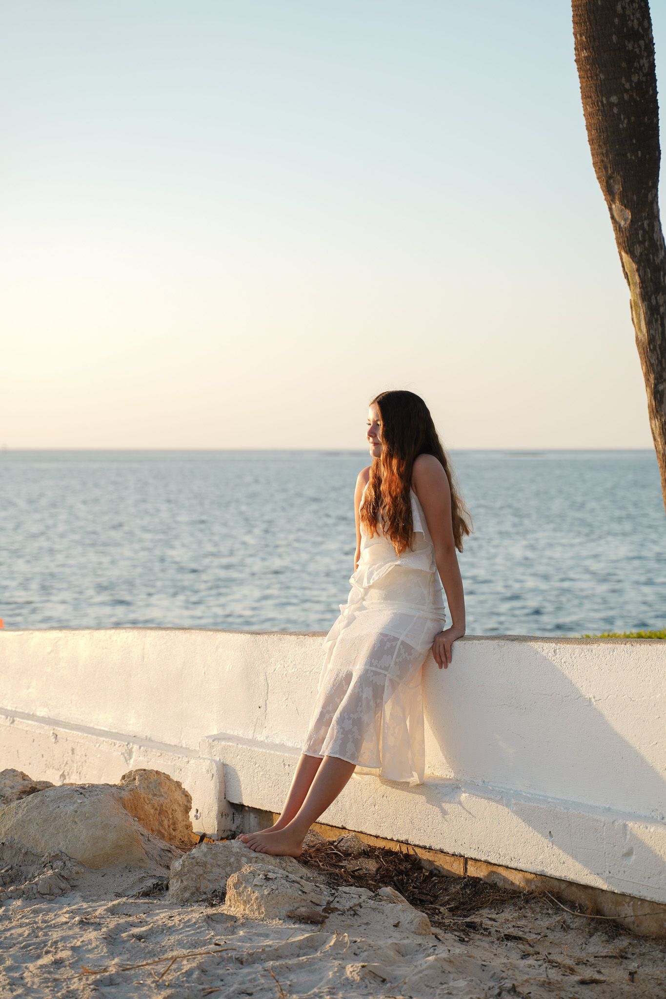 A person in a white dress leans against a wall overlooking the ocean at sunset.