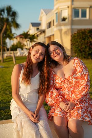 Two people in summer dresses sitting together on a white wall with coastal homes in the background.