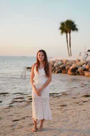 A person in a white dress poses barefoot on a beach with palm trees and rocky shoreline.