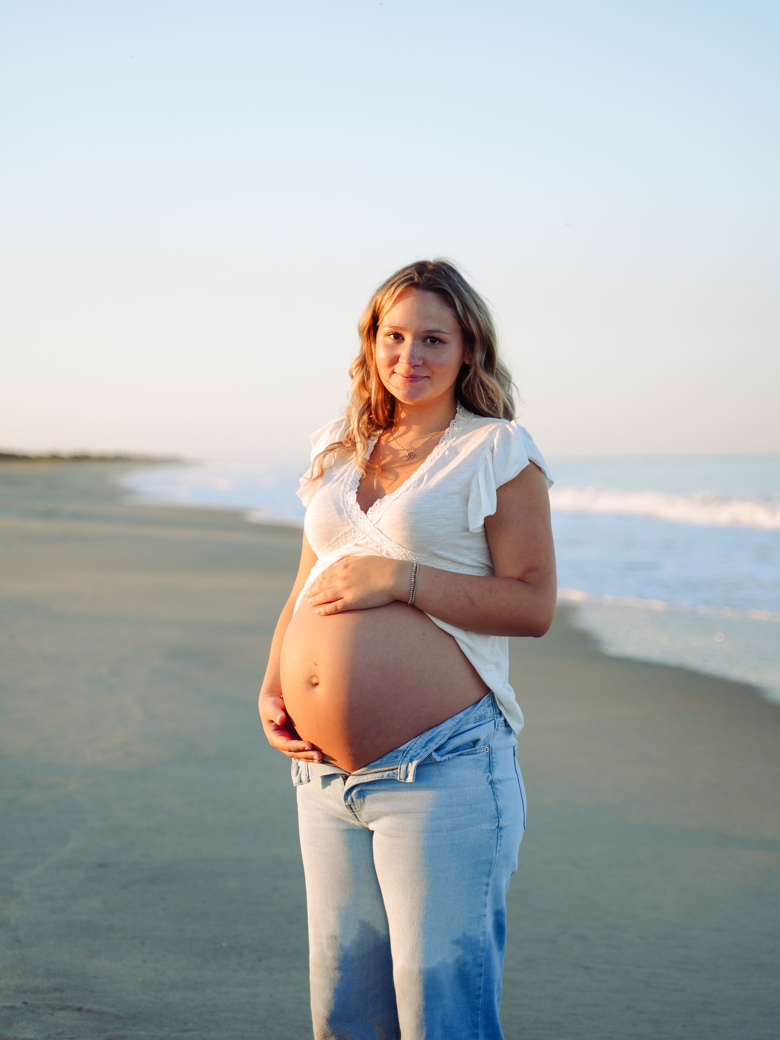 A pregnant woman in a white top and jeans stands on a beach during sunset, cradling her baby bump.
