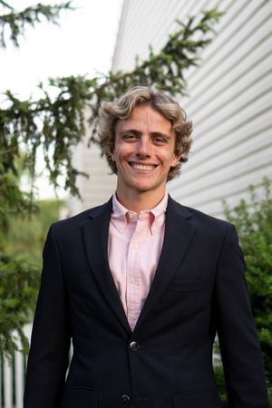 Professional portrait of someone in a dark blazer and pink button-down shirt against white siding and evergreens.