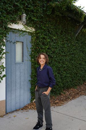 Person in casual navy sweater and gray pants poses against ivy-covered wall with blue door.