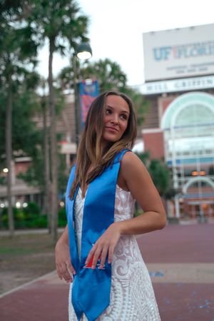 Graduate wearing white dress and blue graduation sash poses in front of UF campus building.