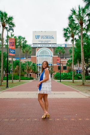 Graduate in white dress and blue sash poses in front of Ben Hill Griffin Stadium entrance.
