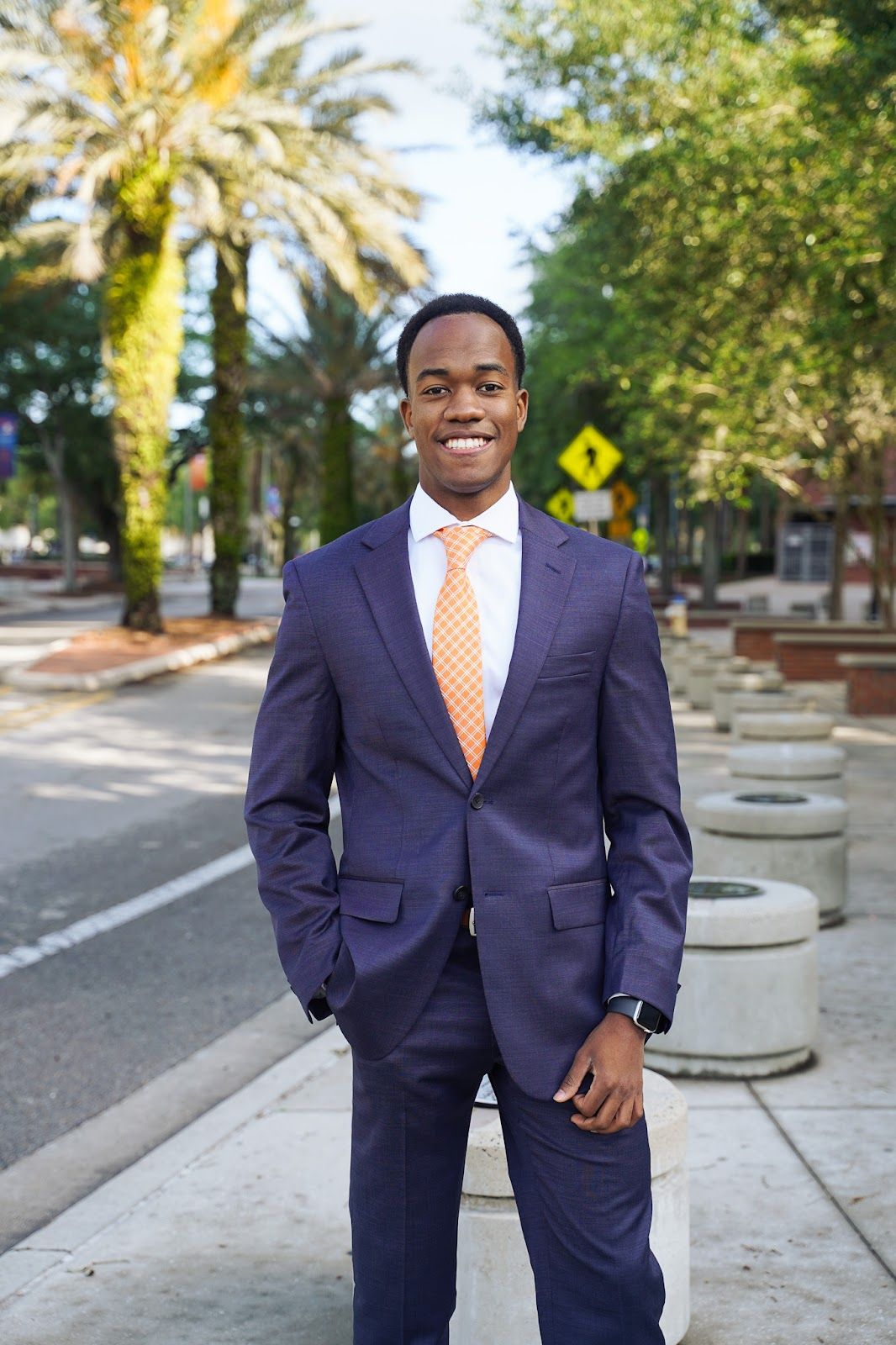 Professional wearing a navy suit with orange tie stands on a palm tree-lined street in business attire.