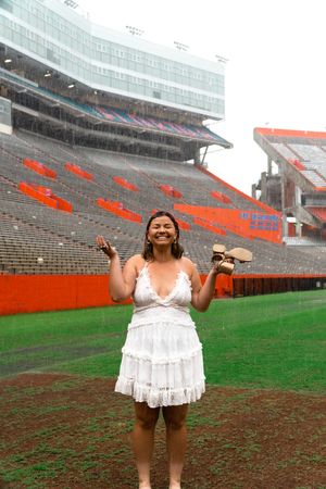 A graduate celebrates on the football field of a stadium wearing a white tiered dress.
