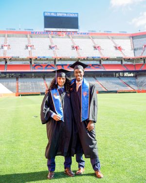 Two graduates in caps and gowns pose on a football field with stadium seating in the background.