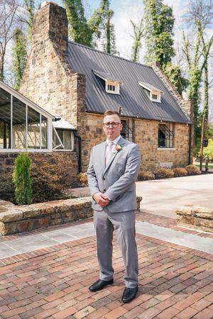 A person in a light gray suit stands on brick pavers in front of a stone cottage with a metal roof and dormer windows.