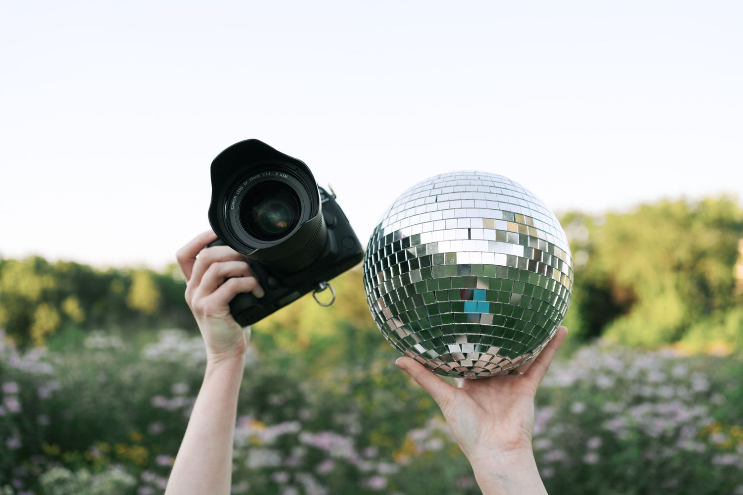 Hands holding up a disco ball and camera against a natural outdoor backdrop.