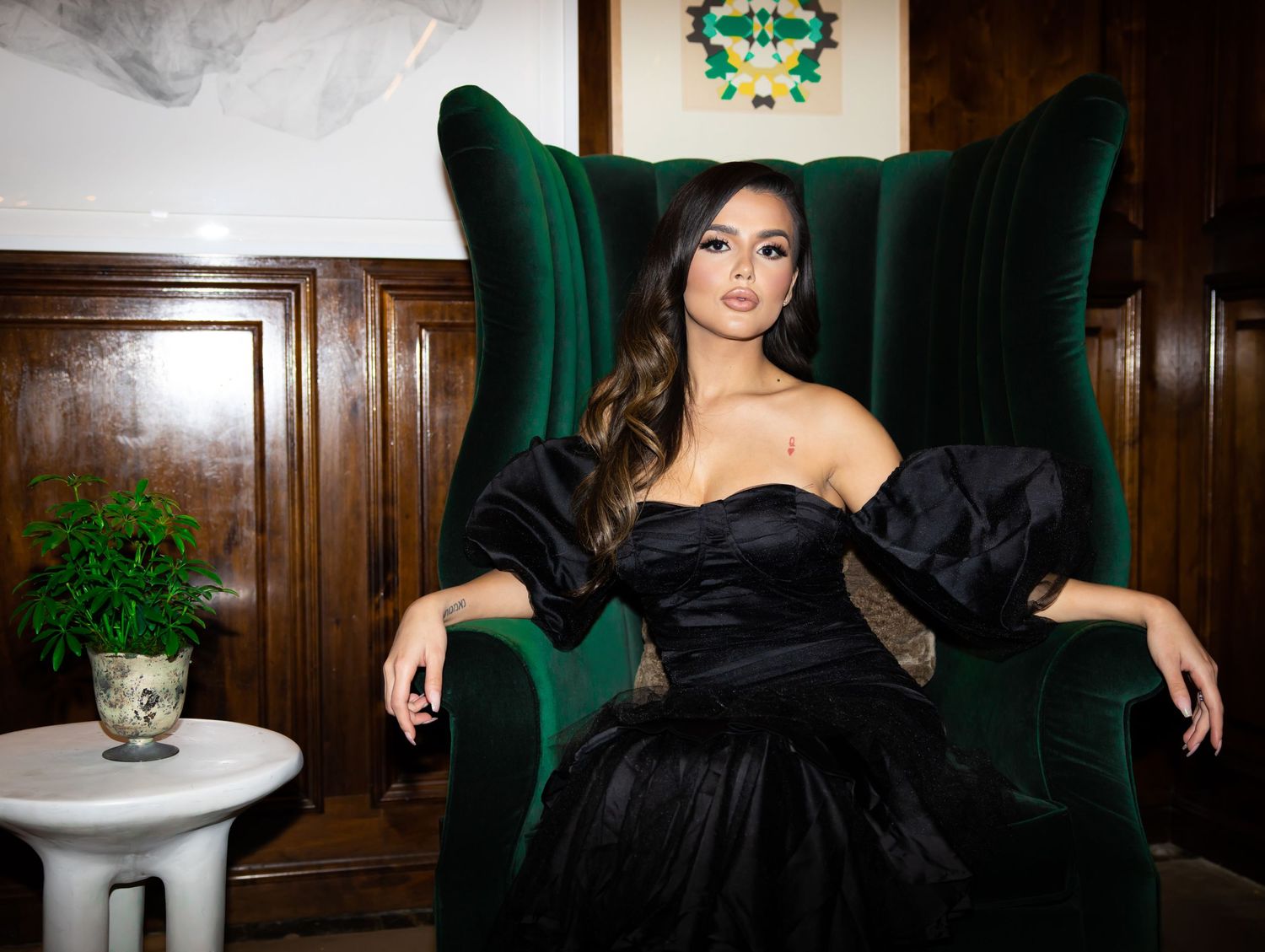 Elegant figure in black off-shoulder gown poses regally in emerald green wingback chair against wood-paneled wall.