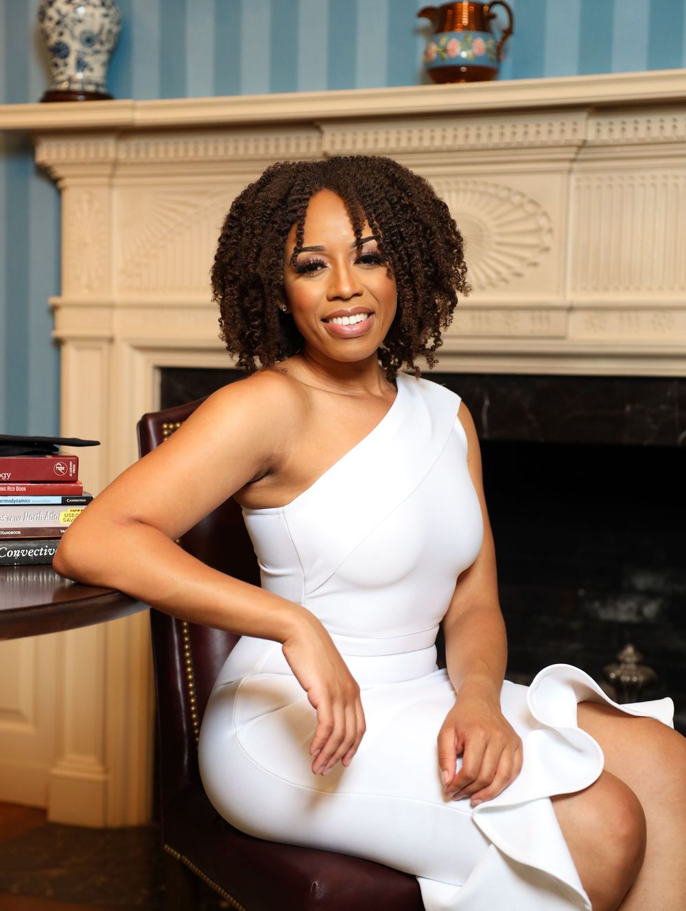 Person in a white one-shoulder dress seated elegantly in a living room with a fireplace and decorative mantle.