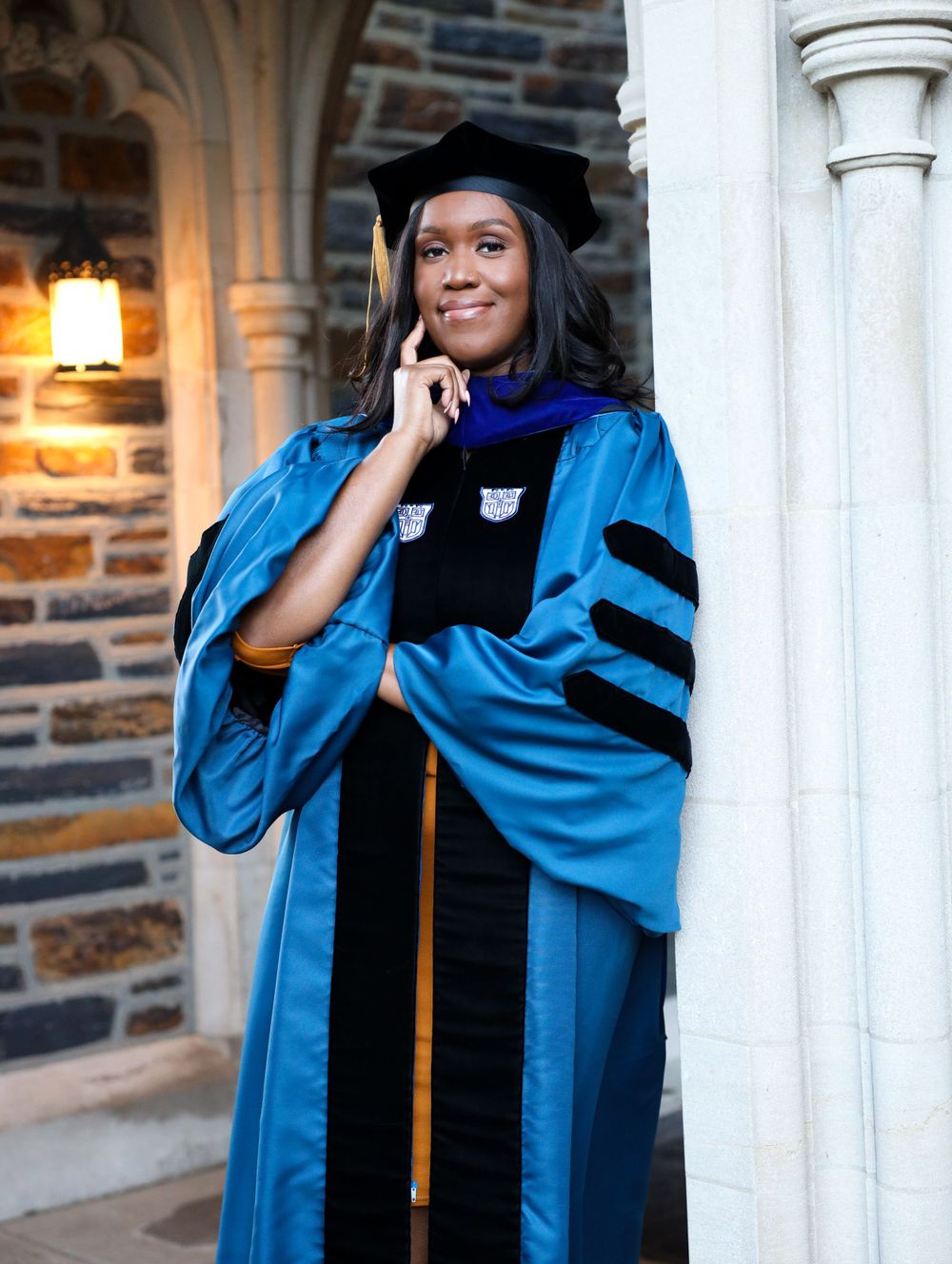 Graduate wearing a bright blue doctoral gown with black velvet trim and gold cord stands smiling by a stone archway.