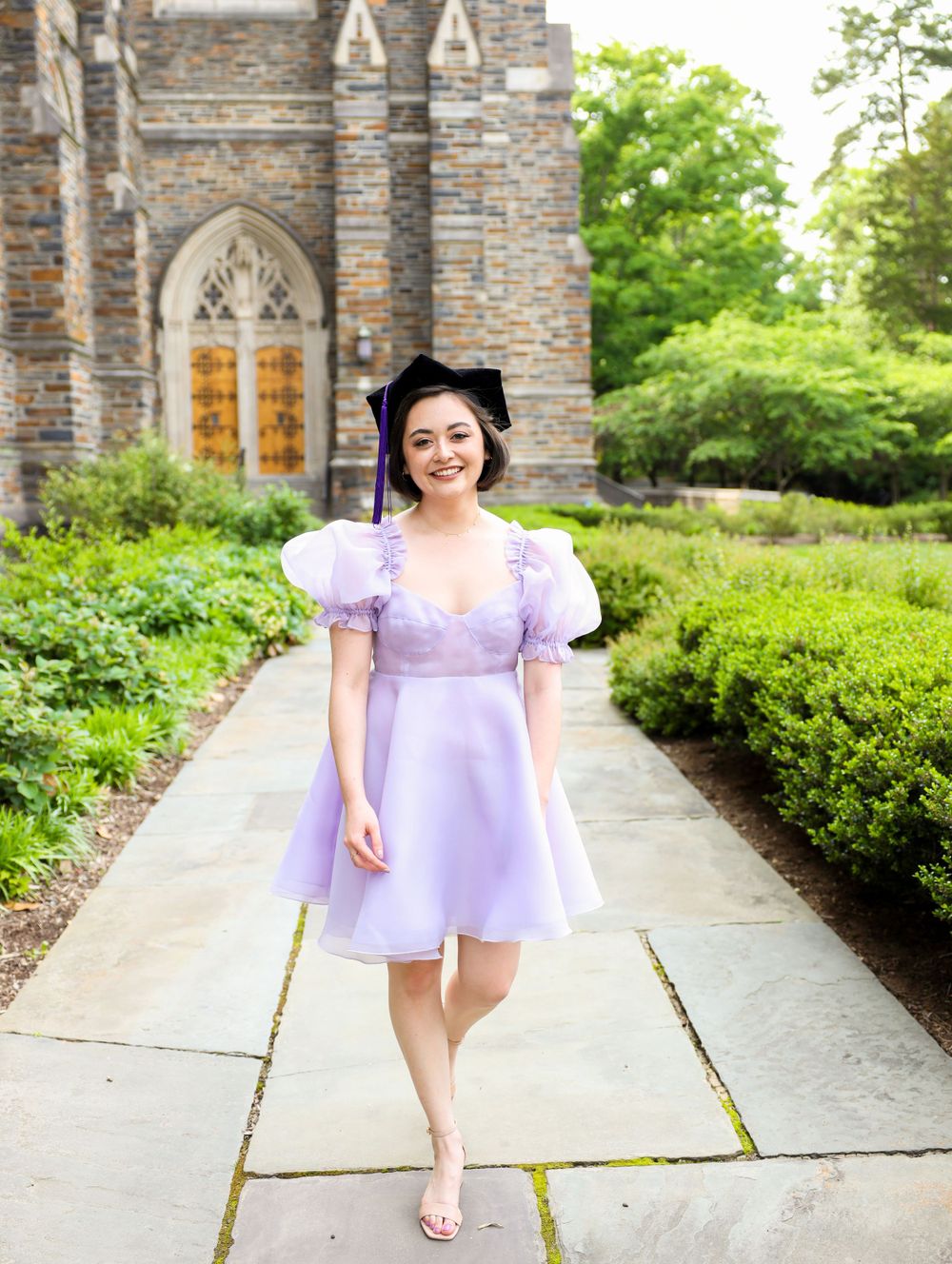 A person in a lilac dress stands on a stone path in front of a gothic stone church with wooden doors.