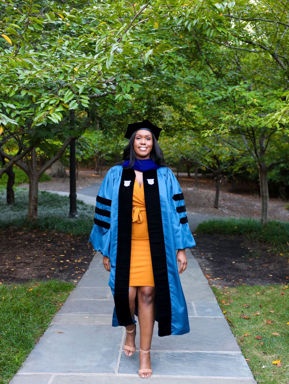 Graduate in blue doctoral regalia and gold dress walks along tree-lined pathway during outdoor graduation celebration.