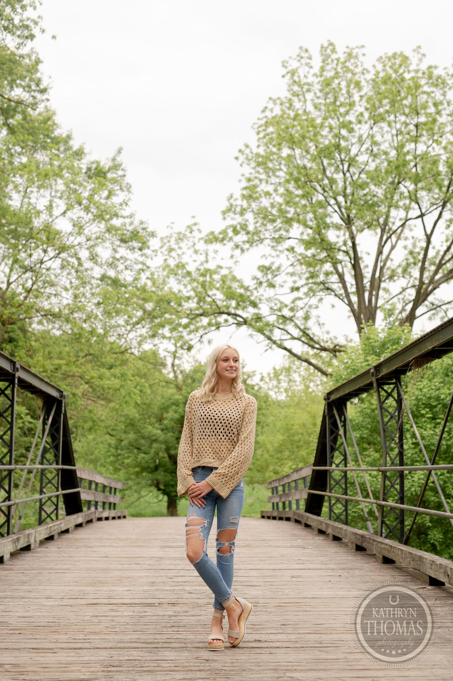 Full-length outdoor fashion photo on wooden bridge with cream sweater and distressed jeans.