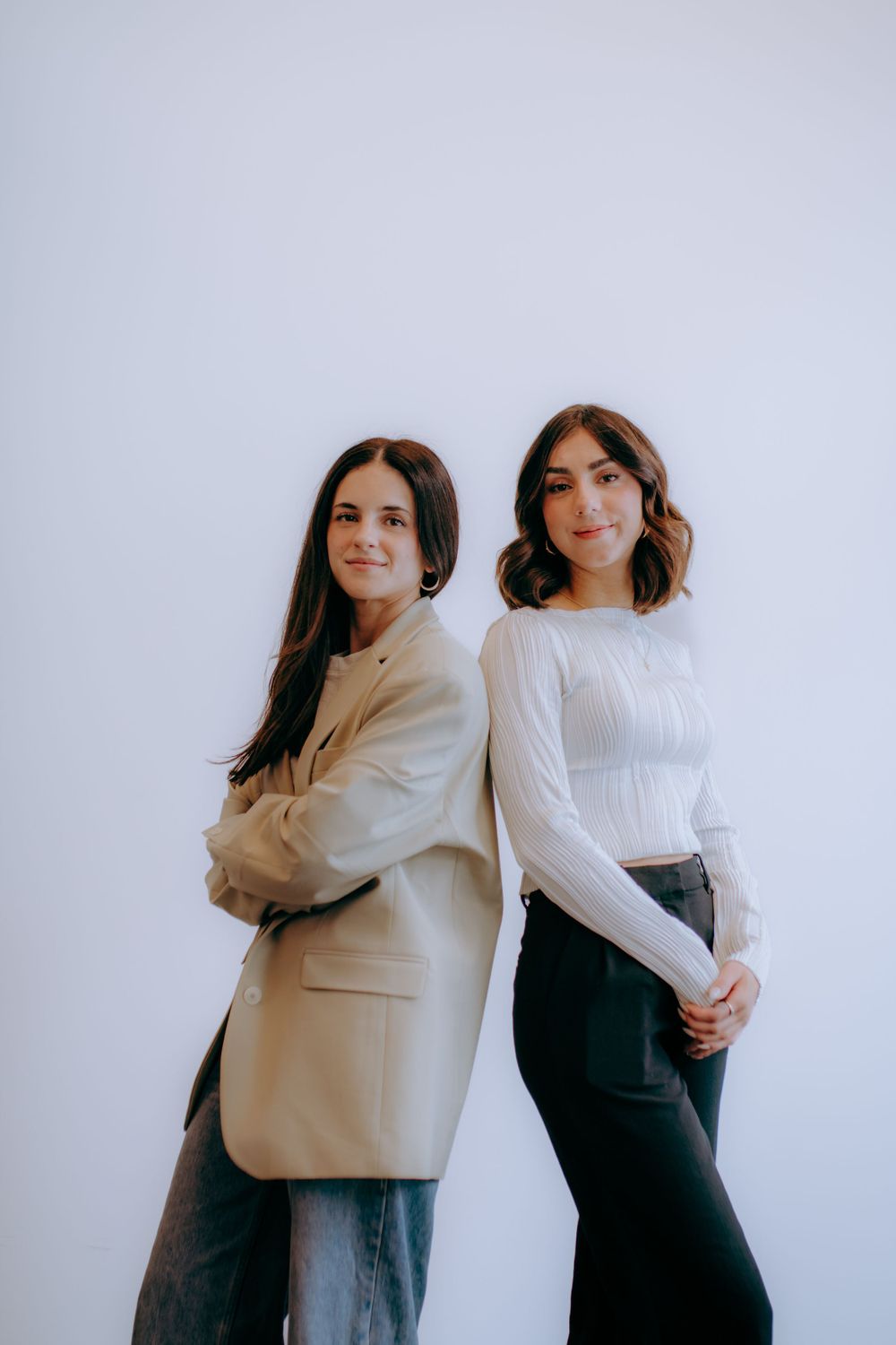 Two women in business casual attire pose against a white wall wearing neutral colors and modern styling.