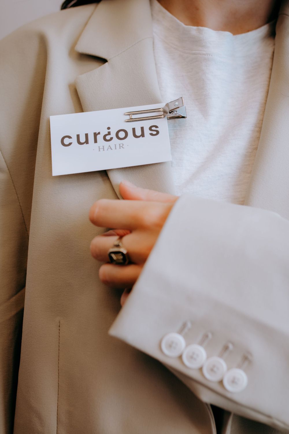 Business card for Curious Hair pinned to a beige blazer with silver ring detail.