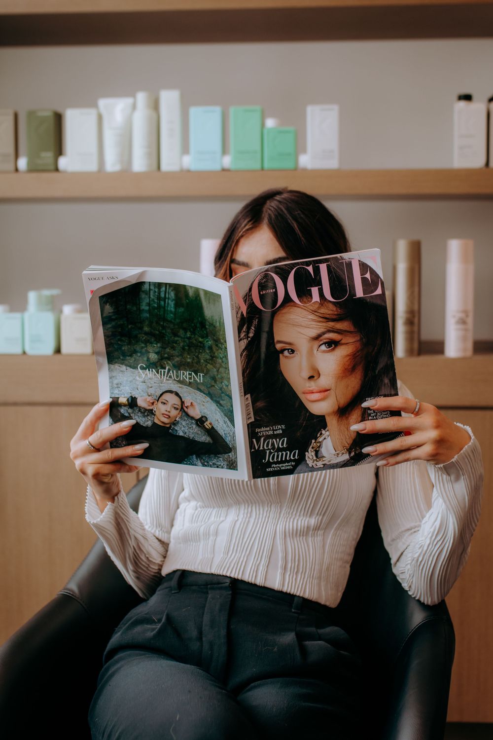 Person reading Vogue magazine in hair salon with product shelves in background.