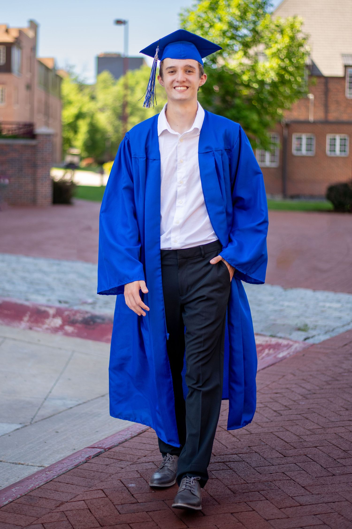 A graduate in traditional blue cap and gown poses on a brick walkway on campus. Cap & Gown mini