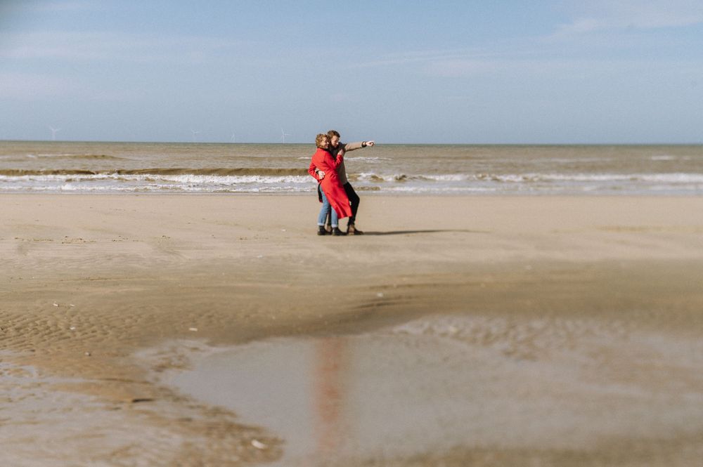 Two figures in red stand together on a vast empty beach with waves in the background.