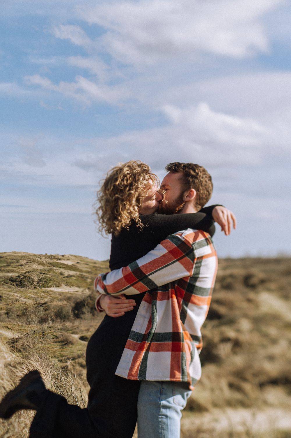 Two people sharing an intimate moment outdoors against a cloudy sky and natural terrain.