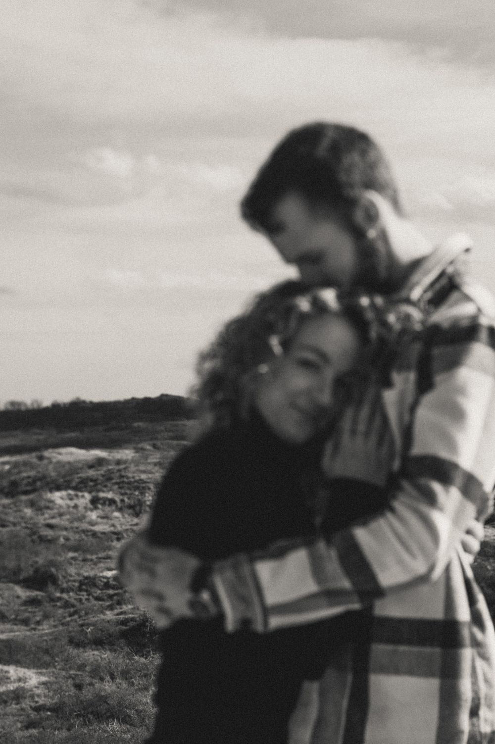 A tender embrace captured in black and white against a cloudy coastal landscape.