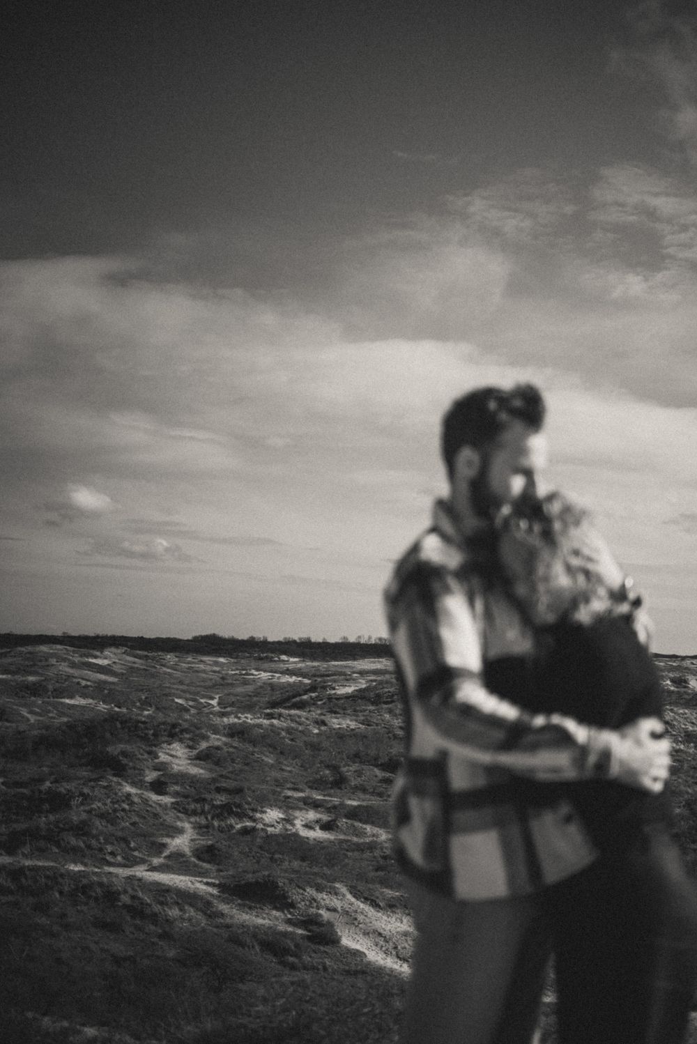 Black and white photograph of two people embracing on a windswept hillside.