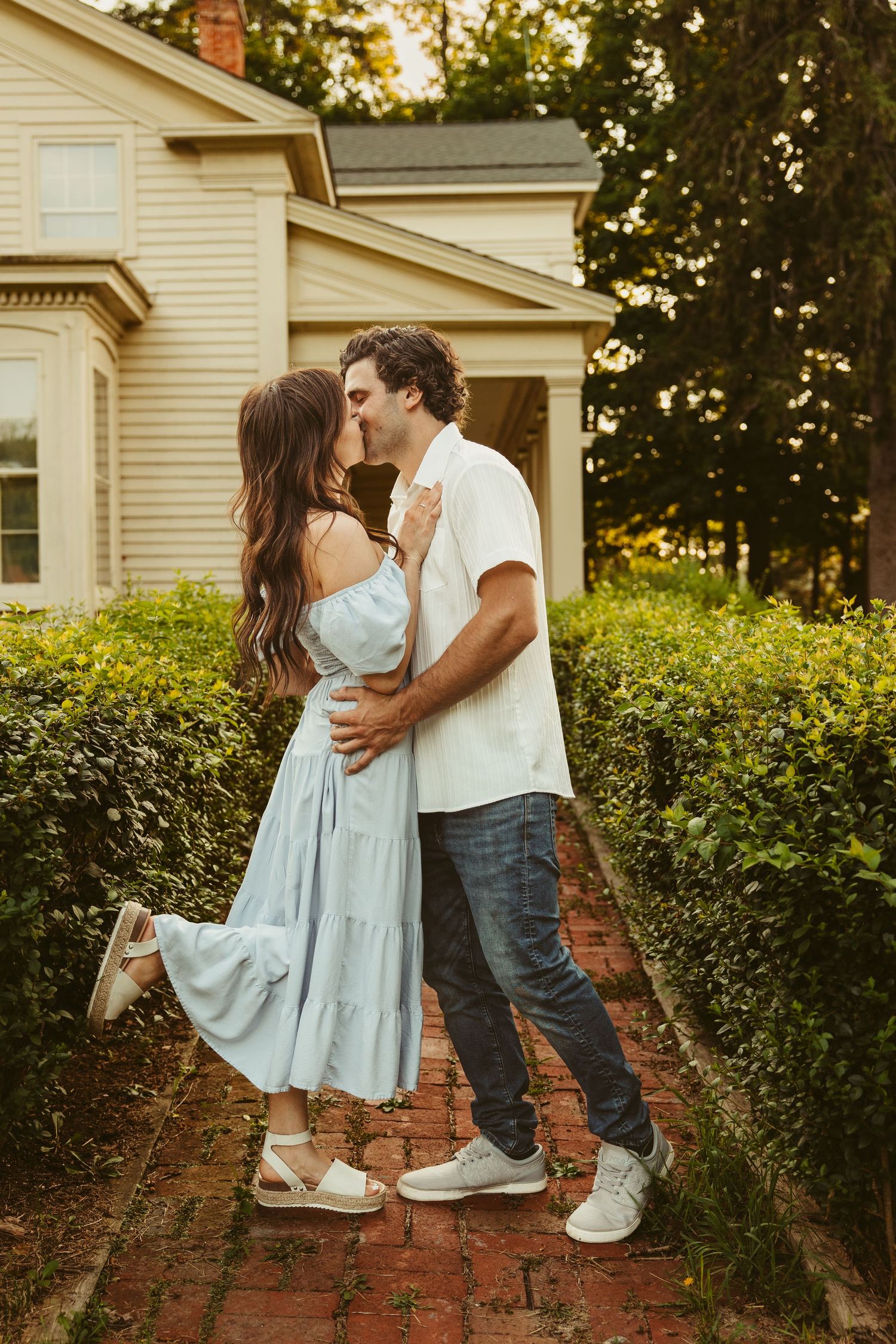A romantic couple embraces on a brick path outside a vintage house during golden hour sunset.