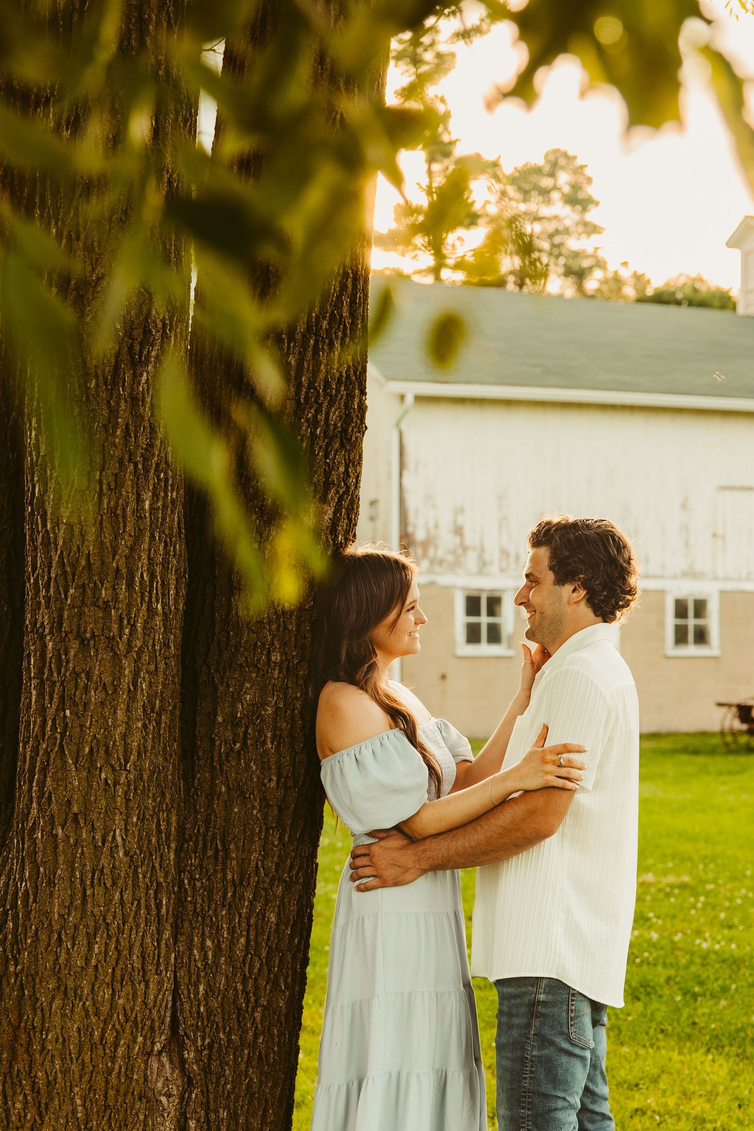 A romantic couple embraces near a tree trunk at golden hour with a white barn in the background.