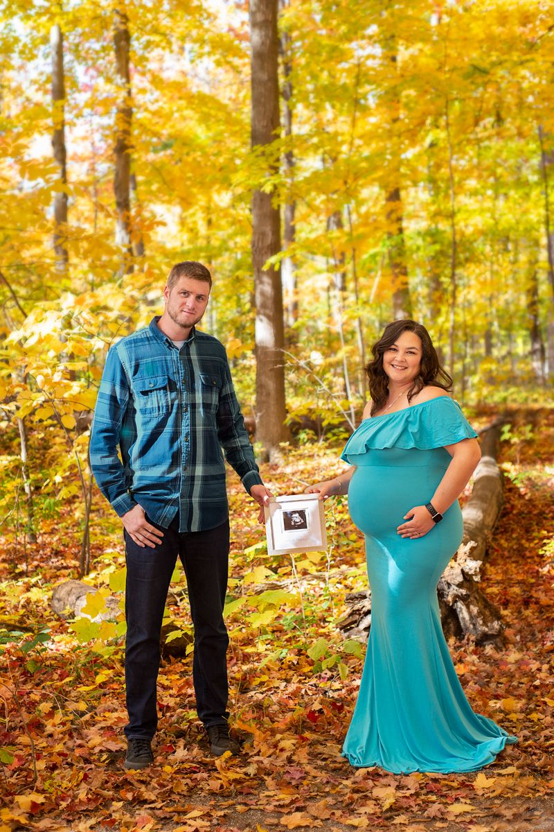 pull back of husband and pregnant wife hold a framed photo of their baby's ultrasound between them in a forrest in the fall