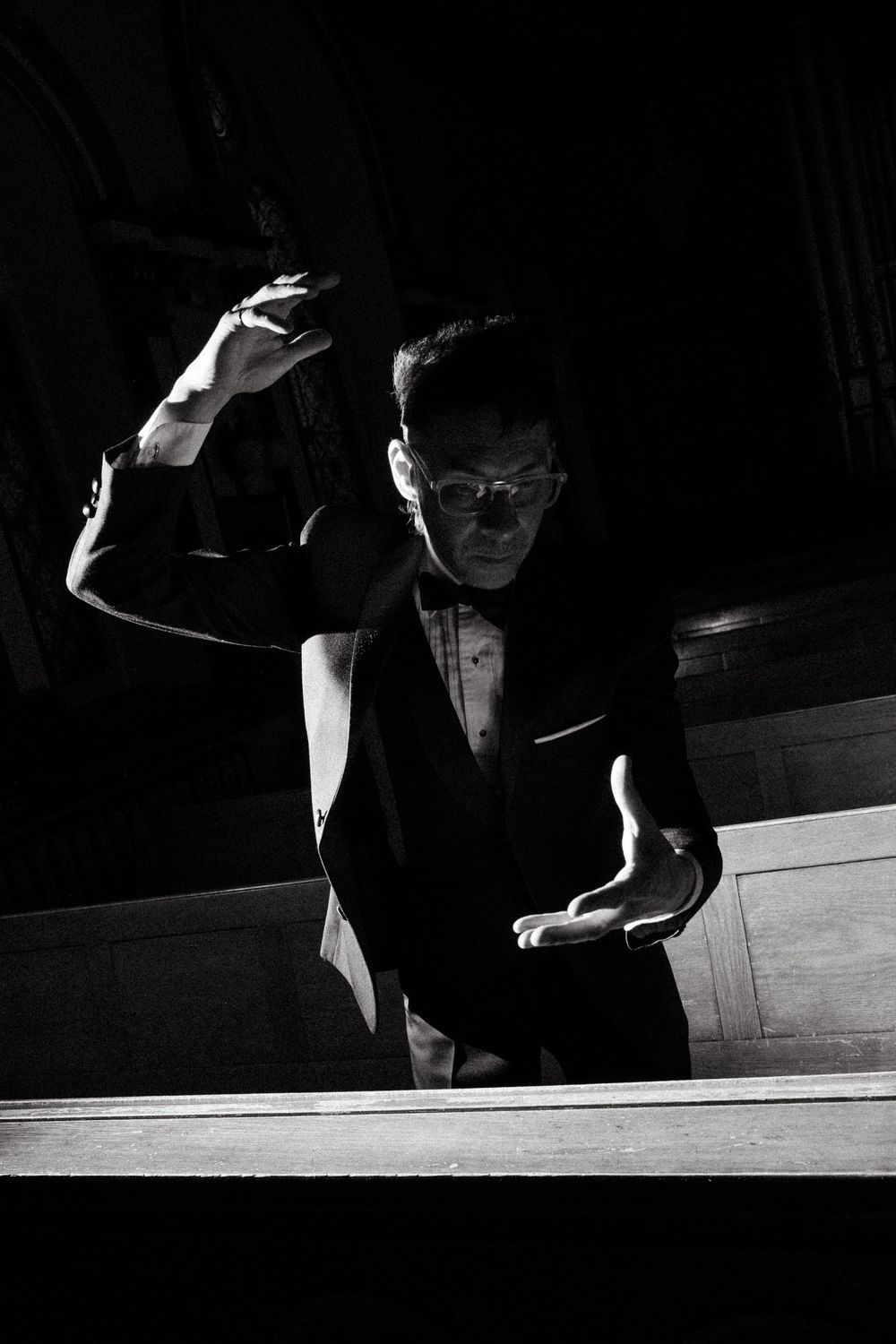 Dramatic black and white photo of a conductor in a tuxedo gesturing expressively during a performance.