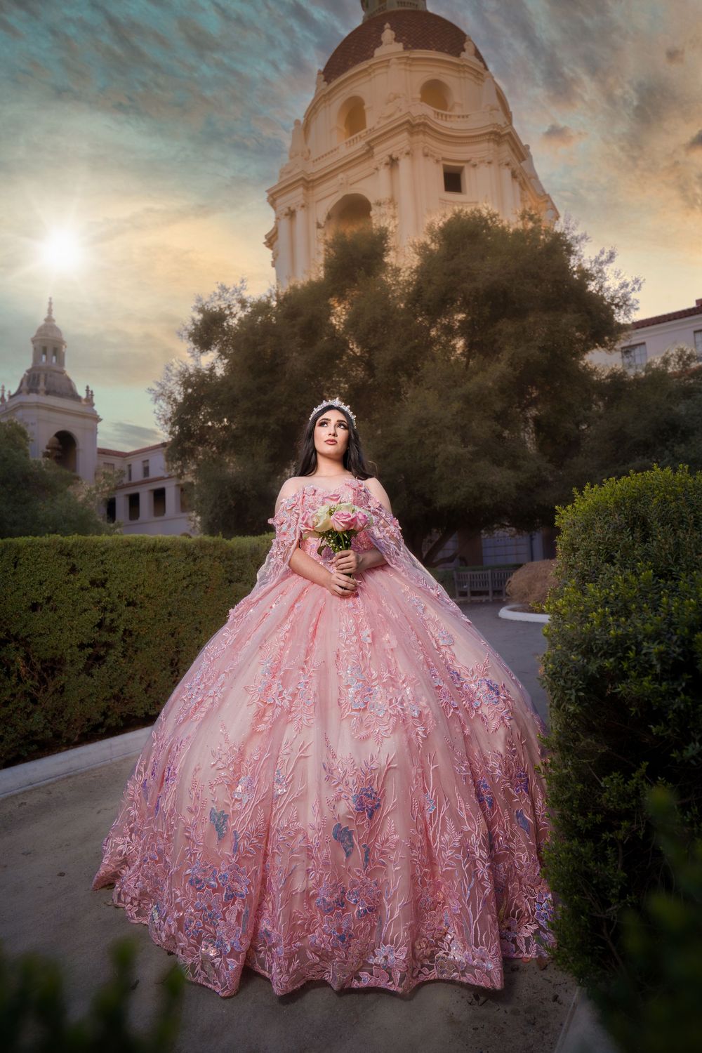 A person in an ornate pink ballgown stands in front of a historic Spanish colonial style building at sunset.