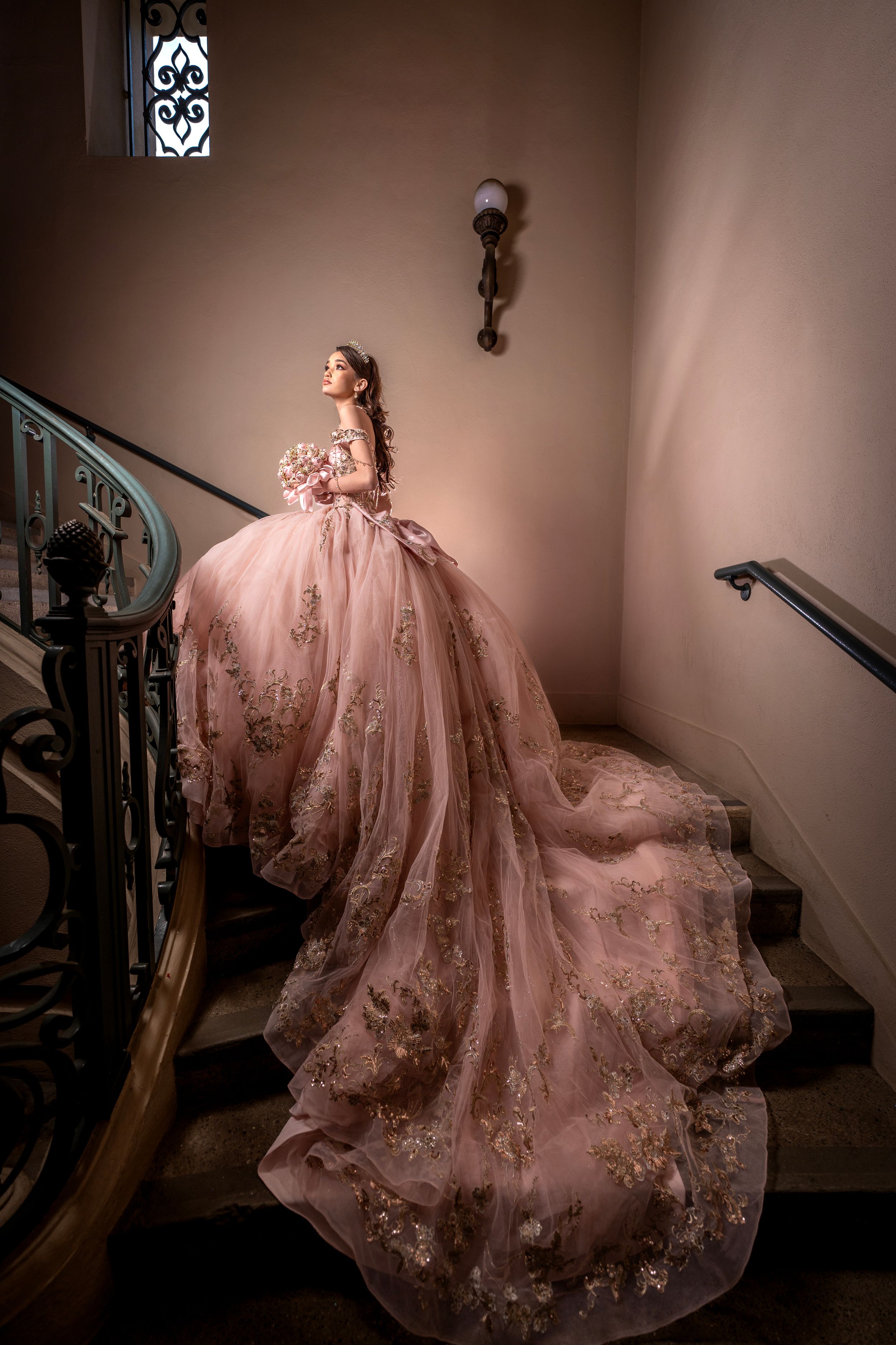 A figure in a flowing blush pink gown with floral appliques poses elegantly on a staircase in dramatic lighting.
