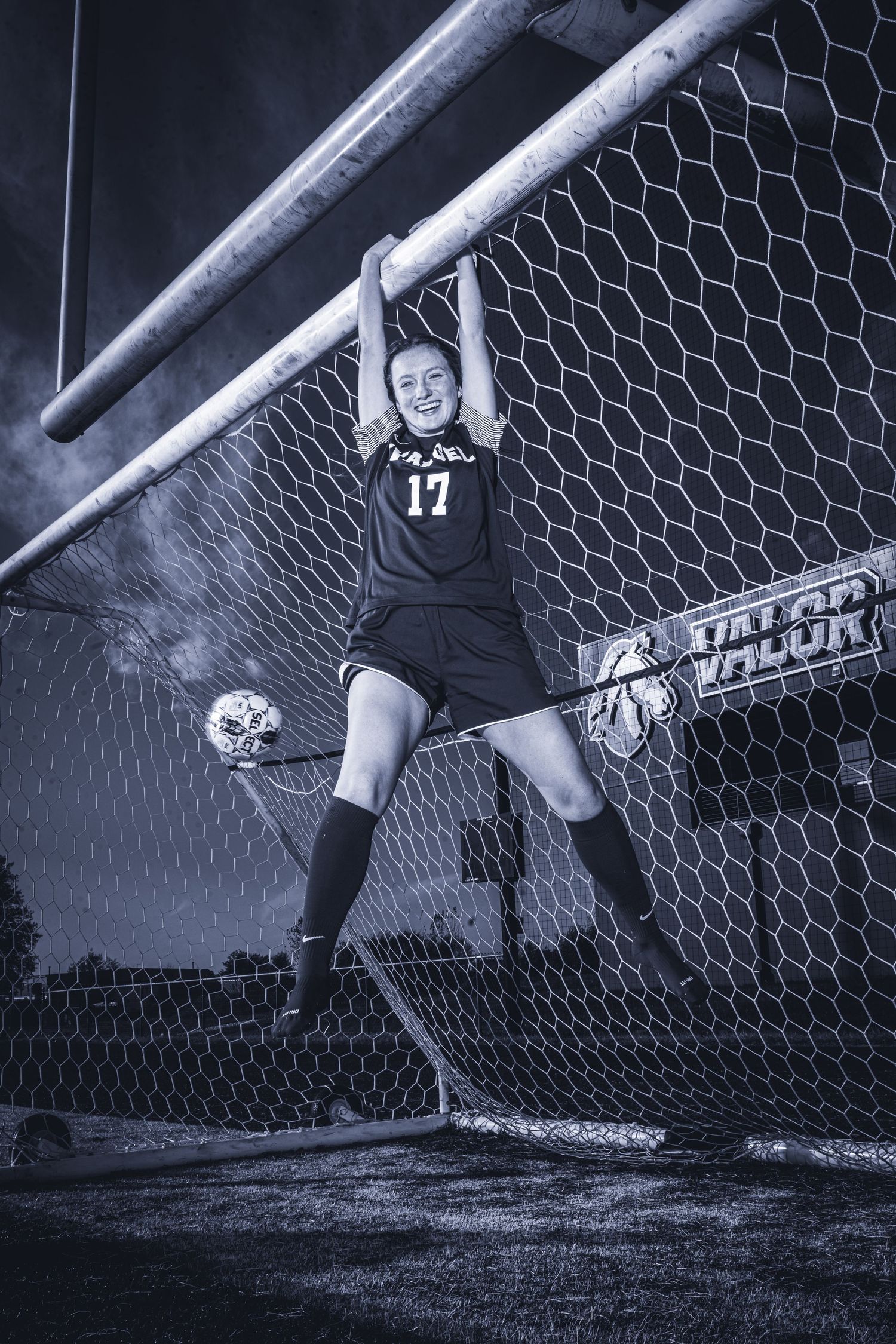 Soccer goalie hanging from crossbar of goal post in dramatic black and white photo.