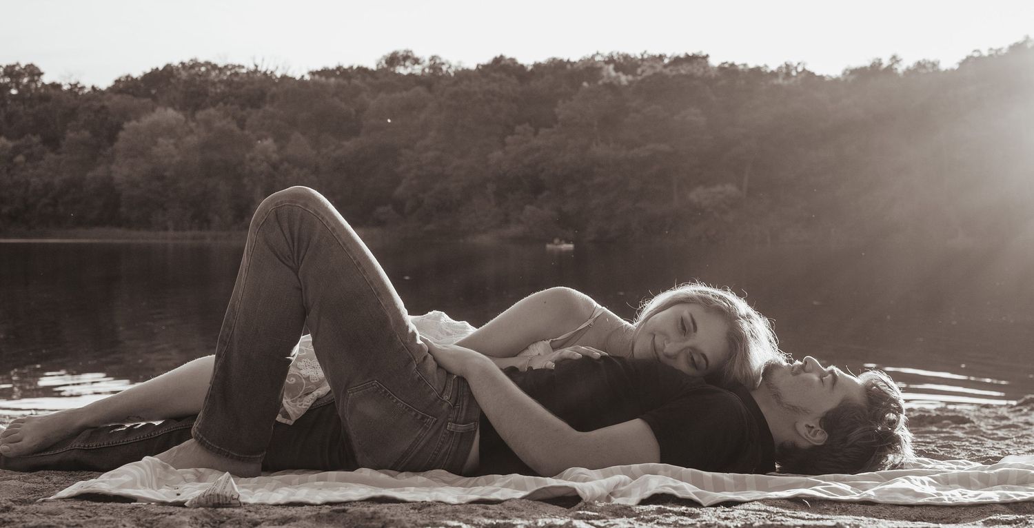 A black and white artistic photograph showing two figures reclining intimately by a calm lake.
